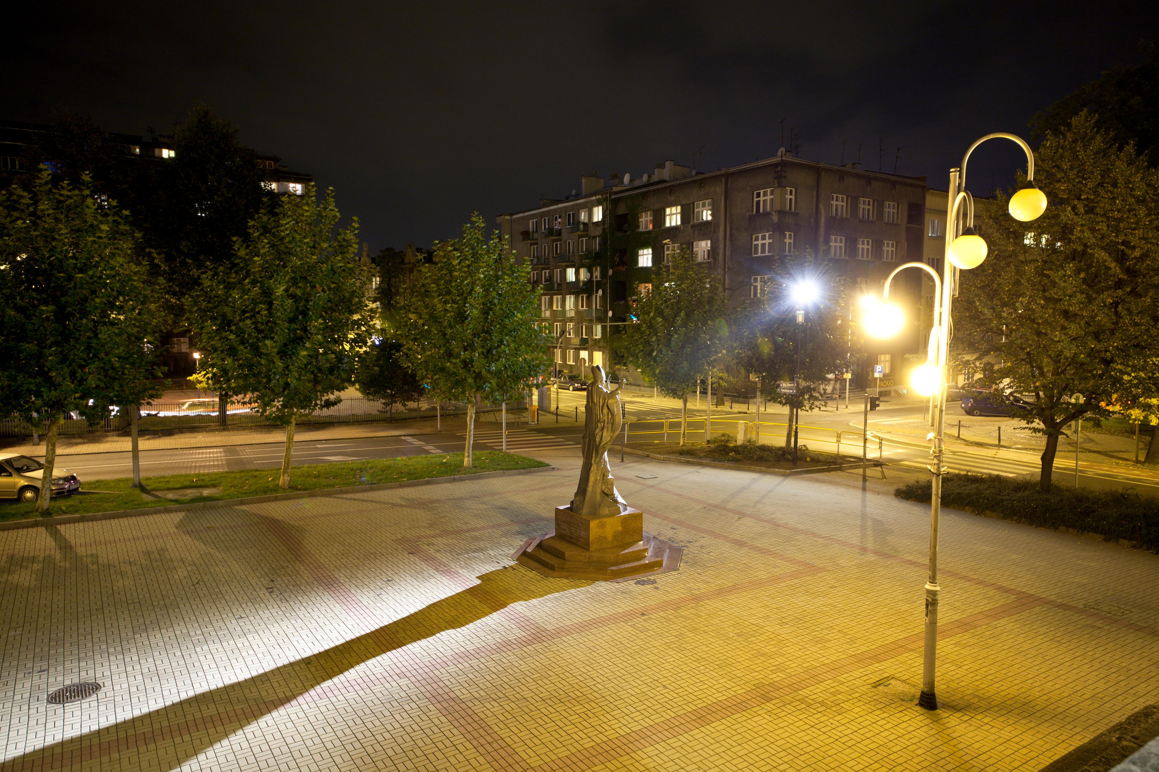 Kilka drzew, lampa oraz mały krzyż na środku małego placu